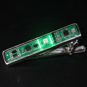 May include: A silver-toned tie clip featuring a rectangular green circuit board inlay. The electronic component surface displays various small black chips, metallic soldering points, and a bright, glowing green LED light integrated into the center.