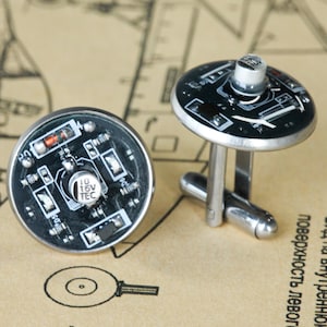 May include: A pair of round cufflinks featuring black circular circuit boards with silver-toned metal hardware. Each cufflink displays electronic components, including a small capacitor labeled 10 16V TEC, resistors, and white printed circuitry lines.