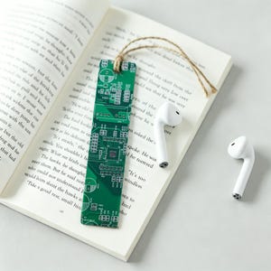 May include: A rectangular bookmark featuring a green printed circuit board pattern with white electronic component diagrams and a brown twine loop at the top, placed on an open book next to two white wireless earbuds.