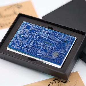 May include: A rectangular metal business card case featuring a blue circuit board pattern on the top surface. The case is presented inside a black foam-lined gift box, showcasing intricate white lines and electronic component designs.
