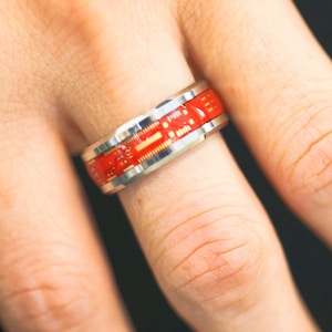 Puede incluir: Un anillo plateado con un diseño de placa de circuito roja. El anillo se lleva en un dedo y presenta un patrón detallado de componentes electrónicos. La banda del anillo es plateada y el diseño de la placa de circuito es rojo y dorado.