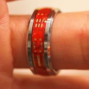 May include: A silver ring with a red and gold circuit board design. The ring is worn on a finger.