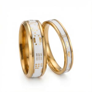 以下が含まれることがあります： 回路基板のデザインが施された白いバンドが付いた、2つのゴールドカラーのリング。リングは研磨仕上げで、モダンな美しさがあります。回路基板のデザインは金色で、白のバンドとのコントラストが際立っています。