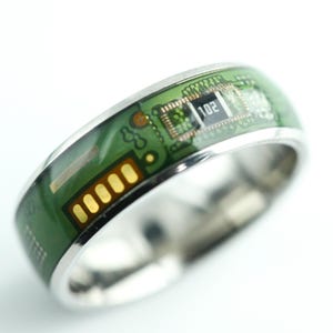 May include: A silver-toned ring featuring a green circuit board inlay. The surface displays intricate gold-coloured electronic traces, a small black rectangular component with the white text 102 printed on it, and a series of five gold contact pads.