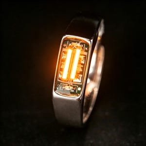 Puede incluir: Un anillo de tono plateado con un componente electrónico rectangular incrustado en el centro. El componente muestra dos barras de luz naranja brillante verticales sobre un fondo de placa de circuito verde oscuro con cableado metálico visible.