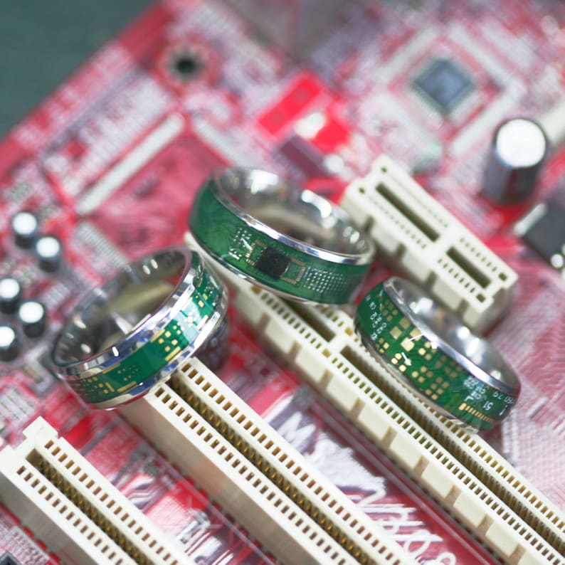 May include: Three silver rings with green circuit board designs on a red and white circuit board. The rings are stacked on top of each other.