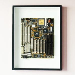May include: Framed vintage computer motherboard with a black frame and white matting. The motherboard is a complex arrangement of chips, connectors, and circuits in shades of gold, green, and black. The IBM logo is visible.