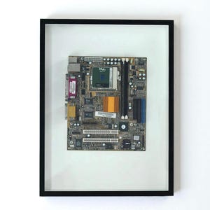 Puede incluir: Placa base de ordenador enmarcada con varios componentes electrónicos. La placa presenta una CPU verde, un disipador de calor naranja y múltiples ranuras y conectores. El marco es negro y el fondo es blanco.
