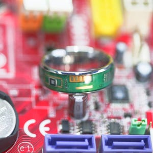 May include: A silver ring with a green and gold circuit board design. The ring is sitting on a red and black circuit board.