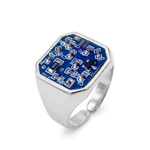 Puede incluir: Un anillo de tono plateado con una cara hexagonal incrustada con un componente de placa de circuito azul. La placa muestra varios componentes electrónicos pequeños, incluyendo resistencias y condensadores, con etiquetas blancas como C26 y R1.