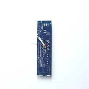 May include: A blue circuit board wall clock with white clock hands and red accents. The clock has the words "DANGER HIGH VOLTAGE" printed on it. The clock is rectangular and mounted on a white background.