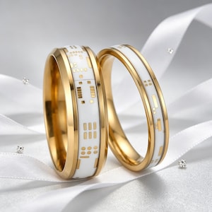 May include: Two gold wedding rings with a white enamel inlay featuring a circuit board design. The rings are displayed on a white ribbon, with small diamond accents. The rings have a polished finish and a modern aesthetic.
