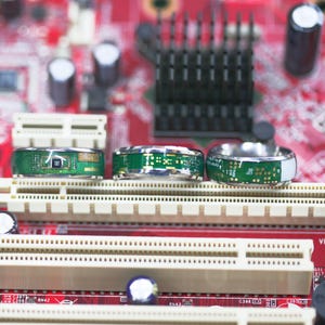 May include: Three silver rings with green circuit board designs. The rings are on a red and white background.