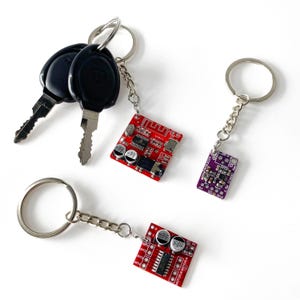 May include: Keychains featuring small red and purple circuit boards with silver key rings. One keychain has a Bluetooth symbol. The keys are black. The circuit boards have various electronic components.