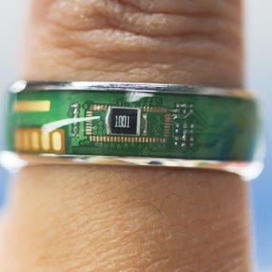 May include: A green circuit board ring with a silver band. The circuit board has a small chip with the number "1001" printed on it.