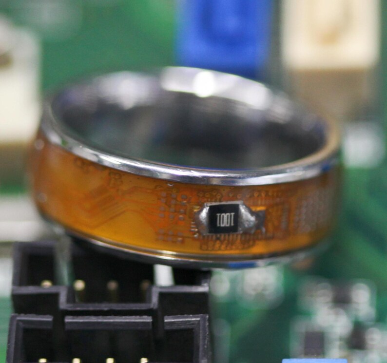 May include: A silver ring with a clear, orange, and silver circuit board design. The ring has the word "TOOT" printed on the circuit board.