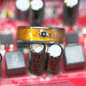May include: Close-up of electronic components on a red circuit board. A gold-colored ring with the text "TOOT" is centered above black cylindrical capacitors and gray rectangular components. The image highlights the intricate details of the circuit board.