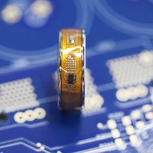 May include: A close-up of a silver and orange ring with a circuit board design. The ring is on a blue surface with a pattern of circles.