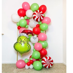 Puede incluir: Un globo verde del Grinch con un gorro rojo de Pap&aacute; Noel, rodeado de una guirnalda de globos rojos, verdes, blancos y rosas. La guirnalda incluye un globo de bast&oacute;n de caramelo.