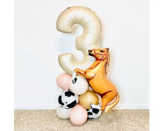 Puede incluir: Un globo de número tres dorado con un globo de caballo marrón unido a él. Los globos están rodeados de globos más pequeños en rosa, blanco y blanco y negro con estampado de vaca.