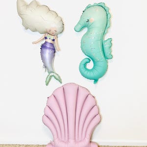 May include: A set of three inflatable decorations featuring a mermaid, a seahorse, and a seashell chair. The mermaid is white with purple and green accents. The seahorse is turquoise with iridescent accents. The seashell chair is pink.
