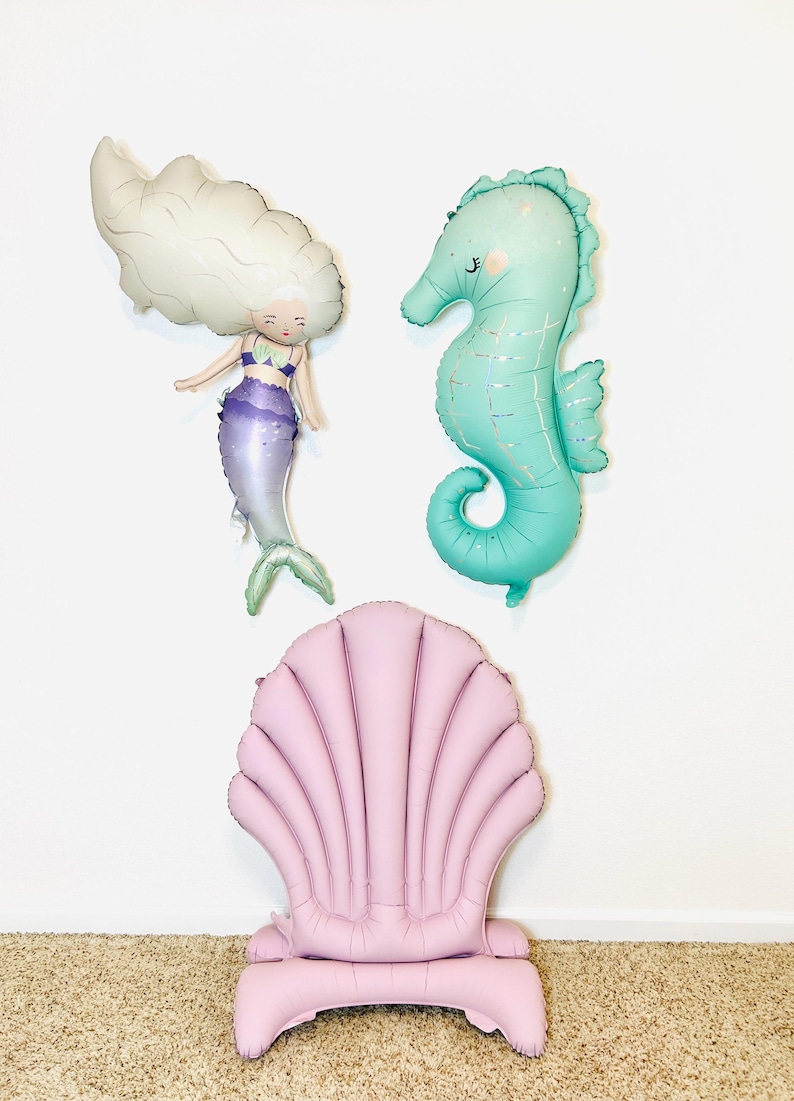 May include: A set of three inflatable decorations featuring a mermaid, a seahorse, and a seashell chair. The mermaid is white with purple and green accents. The seahorse is turquoise with iridescent accents. The seashell chair is pink.