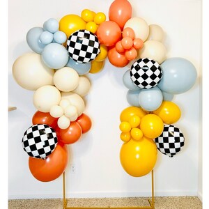 May include: A balloon arch made of white, yellow, orange, blue and black balloons. The balloons are arranged in a semi-circle shape and are held together by a clear string. The balloons are decorated with a chequered pattern.