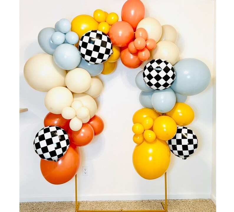 May include: A balloon arch made of white, yellow, orange, blue and black balloons. The balloons are arranged in a semi-circle shape and are held together by a clear string. The balloons are decorated with a chequered pattern.