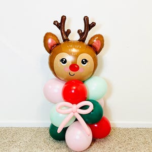 May include: A brown reindeer balloon with a red nose and a pink bow. The balloon is surrounded by green, pink, and red balloons.
