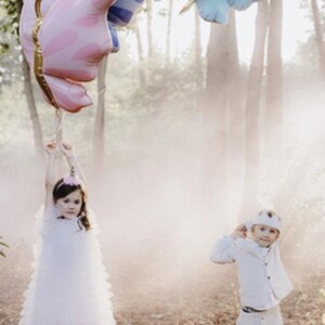 May include: Two children in a forest setting with a light fog. A girl in a white dress holds a pink butterfly balloon. A boy in a white shirt and pants holds a blue butterfly balloon.