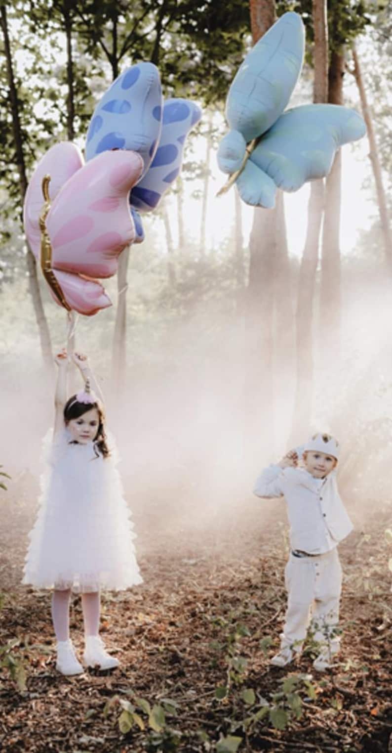 May include: Two children in a forest setting with a light fog. A girl in a white dress holds a pink butterfly balloon. A boy in a white shirt and pants holds a blue butterfly balloon.