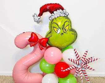 May include: A festive balloon arrangement featuring a Grinch-themed face wearing a Santa hat, a pink number 2 balloon with a red bow, and a cluster of colorful balloons in pink, green, red, and white. A star-shaped decoration adds to the holiday theme.