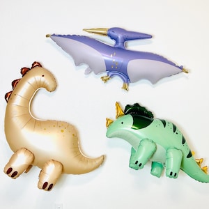 May include: Three inflatable dinosaur balloons: a purple pterodactyl, a brown brontosaurus, and a green triceratops.