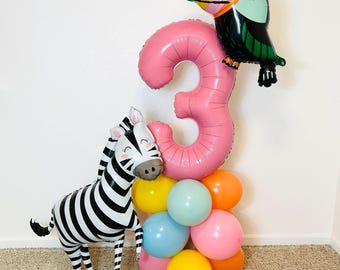 Pode incluir: Decoração de balões para uma festa. Um balão rosa com o número 3, um balão de tucano e uma zebra. O número 3 é suportado por um grupo de balões coloridos em amarelo, azul, rosa e laranja.