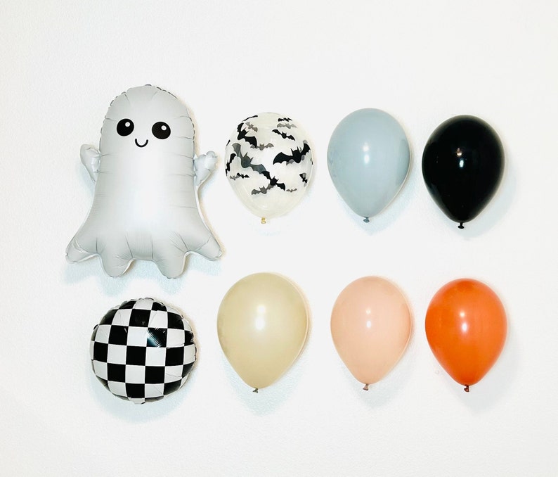 Pu&ograve; includere: Un set di decorazioni per feste di Halloween con un palloncino fantasma argentato, un palloncino a scacchi bianco e nero, un palloncino nero, un palloncino grigio chiaro, un palloncino crema, un palloncino pesca, un palloncino arancione e un palloncino trasparente con un motivo a pipistrello nero.