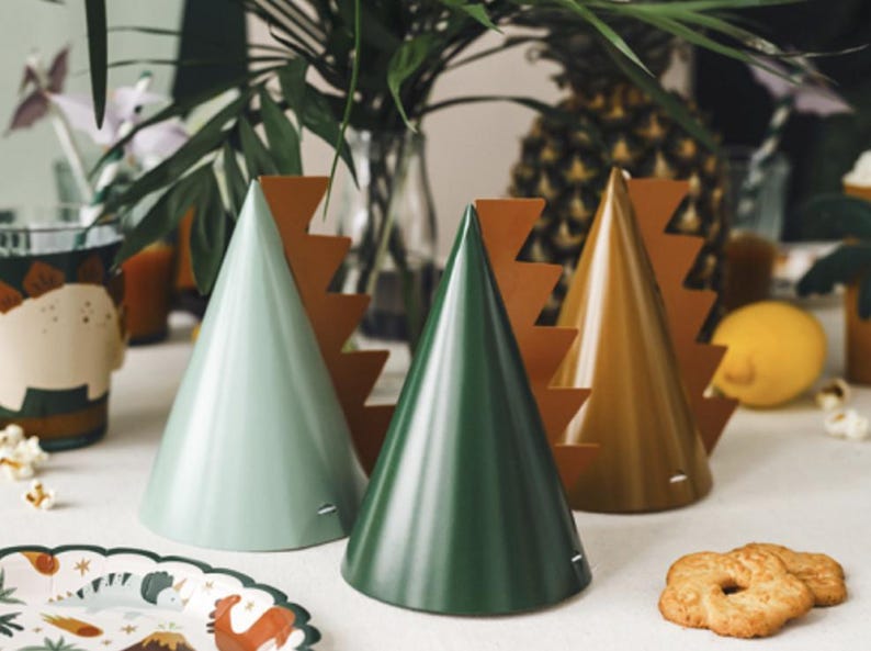 May include: Four party hats with a dinosaur theme. The hats are green, brown, and light green with a brown dinosaur tail design.