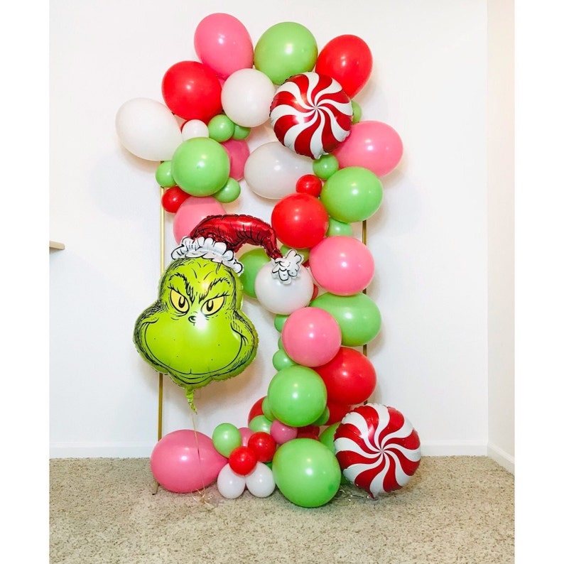 Puede incluir: Un globo verde del Grinch con un gorro rojo de Pap&aacute; Noel, rodeado de una guirnalda de globos rojos, verdes, blancos y rosas. La guirnalda incluye un globo de bast&oacute;n de caramelo.