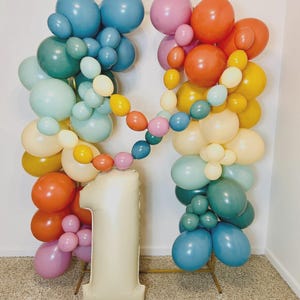 May include: A balloon arch display featuring a mix of matte balloons in shades of blue, green, orange, yellow, pink, and cream. A large, cream-colored foil balloon shaped like the number one stands at the base of a gold metal frame structure.