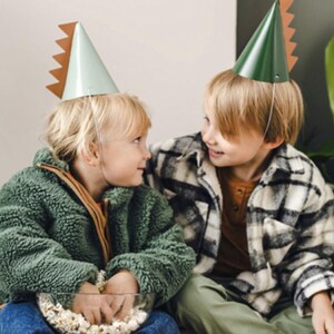 May include: Two children wearing green and white dinosaur party hats. The child on the left is holding a bowl of popcorn. The child on the right is wearing a plaid shirt.