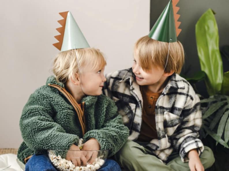 May include: Two children wearing green and white dinosaur party hats. The child on the left is holding a bowl of popcorn. The child on the right is wearing a plaid shirt.