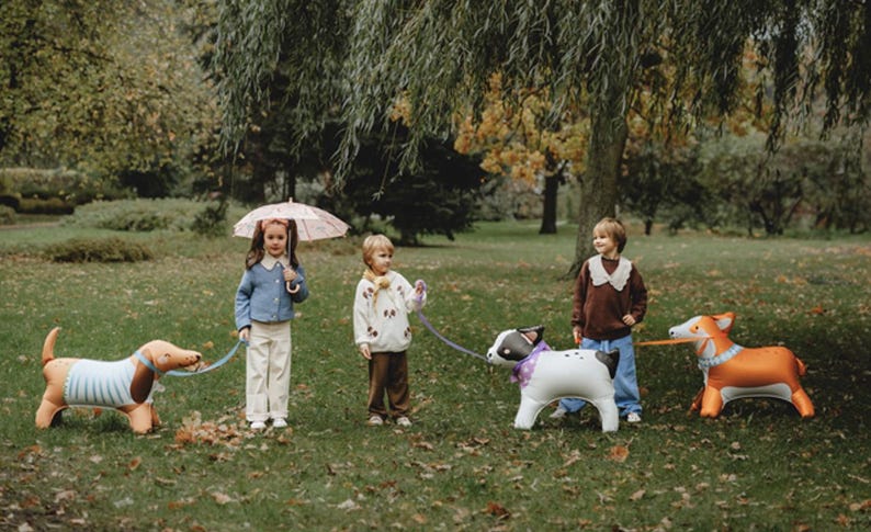 Może przedstawiać: Czterech dzieci bawi się dmuchanymi zabawkami w kształcie ps&oacute;w w parku. Jedno dziecko trzyma r&oacute;żowy parasol. Psy to:  dachshund, corgi i pies w czarno-białe pasy.