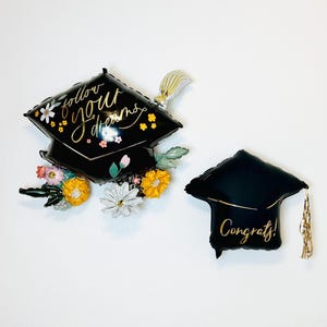 May include: Two black graduation cap shaped balloons. One balloon has the text "Follow your dreams" in gold lettering with floral accents. The other balloon has the text "Congrats!" in gold lettering.
