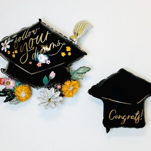 May include: Two black graduation cap shaped balloons. One balloon has the text "follow your dreams" in gold script with floral accents. The other balloon has the text "Congrats!" in gold script.