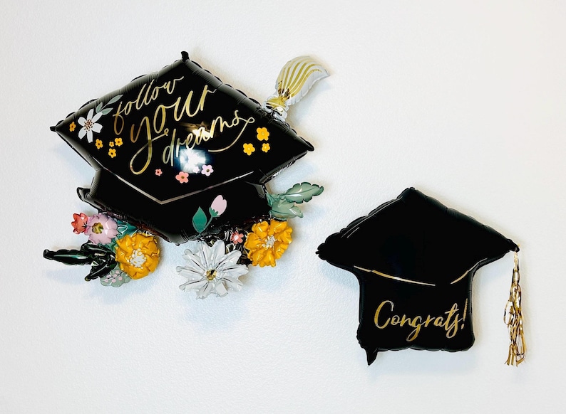 May include: Two black graduation cap shaped balloons. One balloon has the text "follow your dreams" in gold script with floral accents. The other balloon has the text "Congrats!" in gold script.