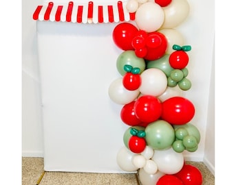 May include: A festive balloon garland with red, white, and green balloons, accented with red and green berry-shaped balloons. The garland is attached to a white backdrop with a red and white striped awning.