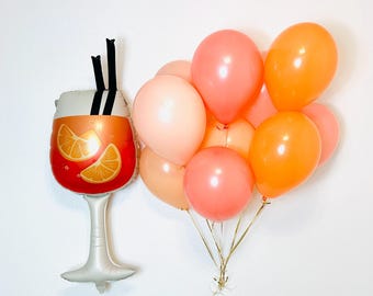 May include: A foil balloon shaped like a cocktail glass with orange liquid and two black straws, accompanied by a cluster of round latex balloons in shades of peach, coral, and bright orange, all tied together with thin gold ribbons.