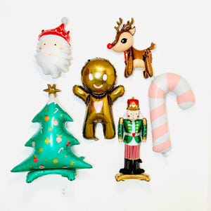 May include: A collection of Christmas-themed foil balloons, including Santa Claus, a reindeer, a gingerbread man, a Christmas tree, a nutcracker, and a candy cane. The balloons are in various colors, such as gold, green, red, and white.