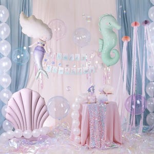 May include: A pink and white mermaid tail balloon, a green seahorse balloon, and a pink seashell balloon are part of a birthday party decoration set. The balloons are surrounded by iridescent balloons and streamers. The text "Happy Birthday" is displayed on a banner.