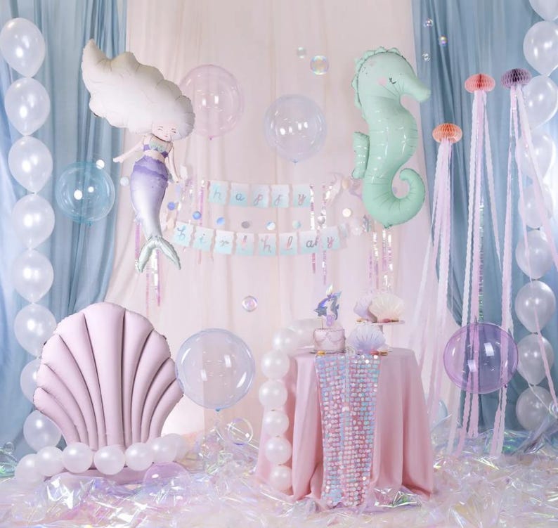 May include: A pink and white mermaid tail balloon, a green seahorse balloon, and a pink seashell balloon are part of a birthday party decoration set. The balloons are surrounded by iridescent balloons and streamers. The text "Happy Birthday" is displayed on a banner.