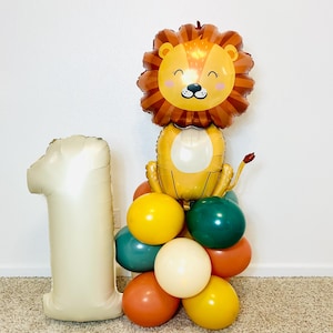 May include: A festive balloon arrangement for a first birthday celebration. It features a large number "1" balloon in a cream color, a lion-shaped balloon, and a cluster of colorful round balloons in shades of yellow, orange, green, and cream.